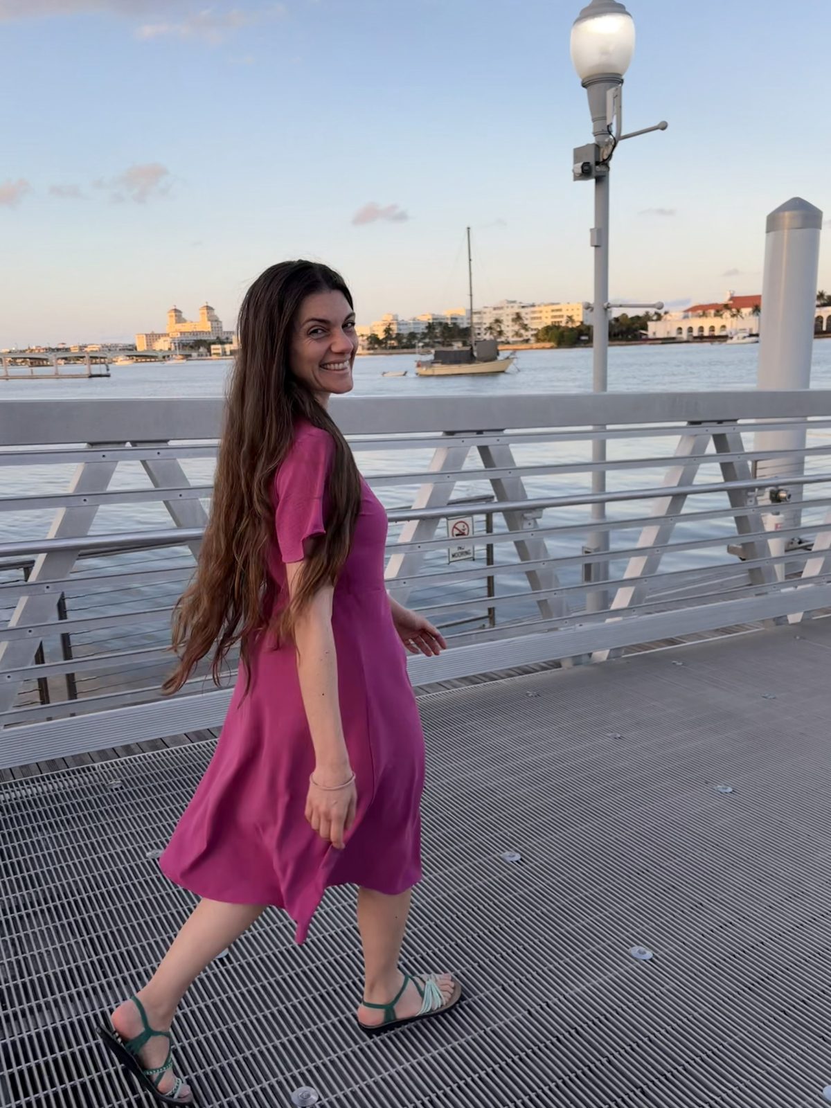 Chiara smiling in a pink dress while walking along a waterfront boardwalk at sunset